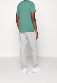 Man som bär en grön t-shirt med kort ärm, ljusgråa träningsbyxor med bakficka, vita strumpor och vita sneakers, som står med ryggen mot kameran.