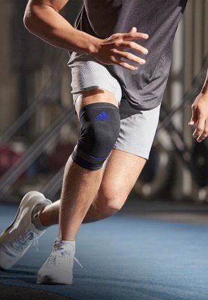 Black athletic knee sleeve with blue Adidas logo, worn over a light gray short. Individual is in motion, wearing white athletic shoes.