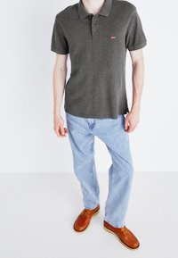 Polo shirt gris foncé avec un col, trois boutons et un petit logo rouge. Porté avec un jean bleu clair et des chaussures à lacets marron.