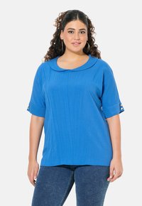 Blusa blu con colletto rotondo, maniche corte, tessuto testurizzato e dettagli con bottoni dorati sulle maniche. Abbinata a jeans scuri.