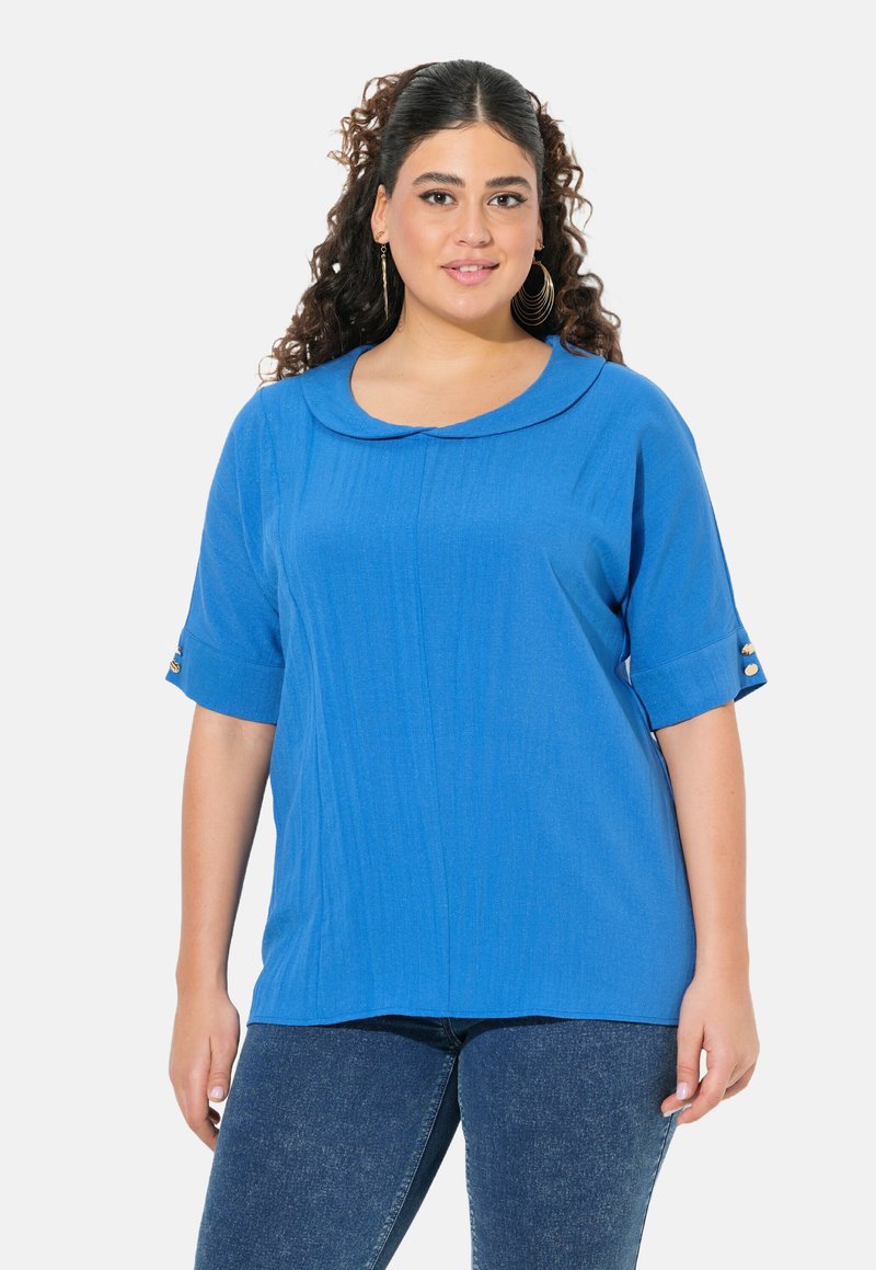 Blusa blu con colletto rotondo, maniche corte, tessuto testurizzato e dettagli con bottoni dorati sulle maniche. Abbinata a jeans scuri.