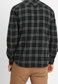 Camisa de franela de cuadros en negro y gris, con mangas largas, cuello abotonado y bolsillo en el pecho, exhibida desde la parte posterior.