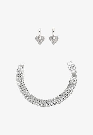 Ezüst karkötő összekapcsolódó láncszemekkel és gyémánt díszítéssel, ezüst, szív alakú gyémánt karika fülbevalókkal fehér háttéren.