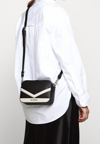 Černá kožená crossbody taška s bílými akcentními pruhy. Disponuje texturovaným povrchem, nastavitelným popruhem a kovovými prvky. Logo na přední straně.