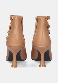 Botas de tobillo de cuero color bronce con un tacón alto, que presentan una parte trasera curvada, doble cremallera y acentos de botones circulares a los lados.