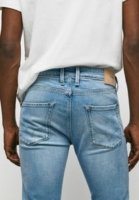 Light blue denim jeans with a fitted design, five pockets, and a visible leather patch at the waist. Smooth texture with subtle fading.
