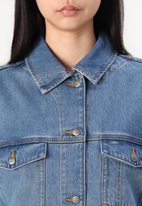 Veste en denim bleu avec un col classique, deux poches poitrine avec des boutons-pression en métal argenté, et des détails de couture orange.