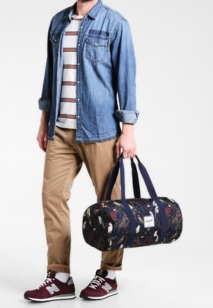 Bolso de deporte con un patrón floral oscuro, forma redondeada, asas de color marino y detalle de cremallera. Llevado con una camisa de mezclilla y pantalones caqui.