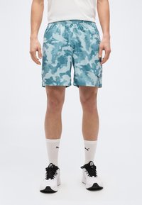 Άνδρας που φοράει μπλε καμουφλάζ αθλητικές βερμούδες, λευκές κάλτσες Puma και λευκά αθλητικά παπούτσια με μαύρα κορδόνια, στεκόμενος μπροστά σε απλό φόντο.