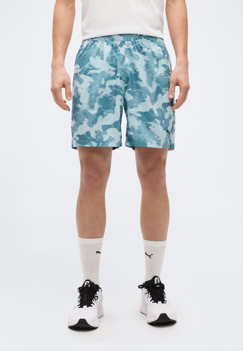 Άνδρας που φοράει μπλε καμουφλάζ αθλητικές βερμούδες, λευκές κάλτσες Puma και λευκά αθλητικά παπούτσια με μαύρα κορδόνια, στεκόμενος μπροστά σε απλό φόντο.