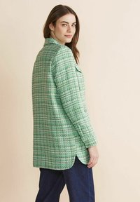 Blazer de tweed verde con un acabado texturizado, bolsillos frontales y un dobladillo con abertura lateral, combinado con pantalones de mezclilla oscuros. Modelo girado ligeramente.