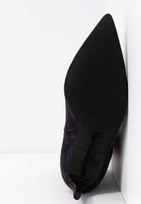 Chaussure à talon haut en suede noir avec un bout pointu et une texture lisse, présentant une silhouette élancée et courbée ainsi qu'une semelle noire unie.