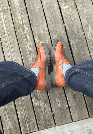 Brune lædersko med teksturerede detaljer og gummisåler, kombineret med blå stribede sokker og denimjeans, placeret på træterrasse.