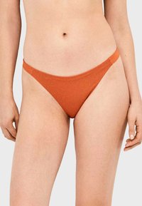 Femme portant un bas de bikini côtelé orange, debout avec les bras détendus le long du corps devant un fond uni.