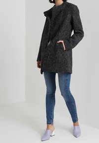 Person klädd i en mörkgrå texturerad kappa, matchande tröja, blå skinny jeans och lila klackskor mot en neutral bakgrund.