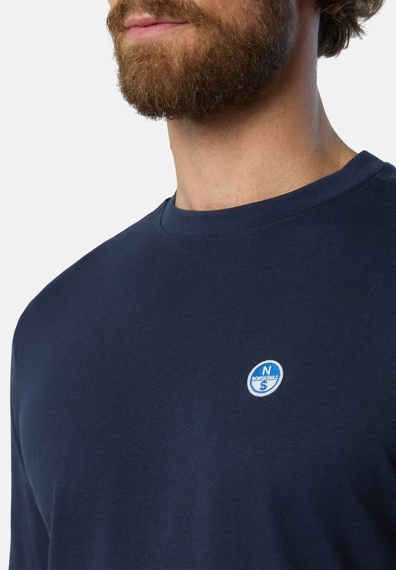 Marinblå långärmad t-shirt i bomull med rund halsringning och en liten cirkulär blåvit logotyp på bröstet. Slät textur.