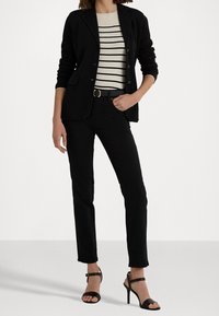 Černý blazer přes pruhovaný smetanový a černý top, doplněný černými slim-fit džínami a černými páskovými podpatky, s páskem s zlatými akcenty.