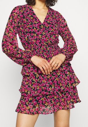 Vrouw die een zwarte jurk met lange mouwen draagt met een roze en bruine bloemenprint en een gelaagde ruchesrok, handen ineen gestrengeld op de taille met ringen.