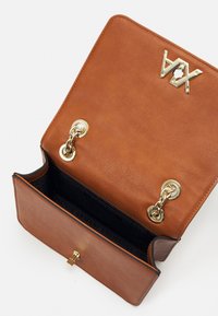 Hnědá kožená kabelka se zlatými kovy. Má zapínání na klopu, řetízkové doplňky a černou podšívku s názvem značky.