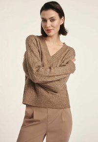 Sweter w sploty kablowe, w dekolcie w kształcie V w jasnobrązowym kolorze, z prążkowanym wykończeniem u dołu i na mankietach; teksturowany wzór w całej długości. W zestawie ze dopasowanymi beżowymi spodniami.