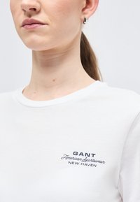 Κοντινό πλάνο ενός ατόμου που φοράει λευκό t-shirt GANT με το κείμενο «American Sportswear New Haven» στο στήθος και ένα μικρό κρίκο σκουλαρίκι.