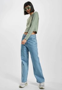 Lys grønn langermet cropped topp med frynset kant, sammen med vide lyseblå jeans og grå sneakers. Modellen har på seg overdimensjonerte grønne solbriller.