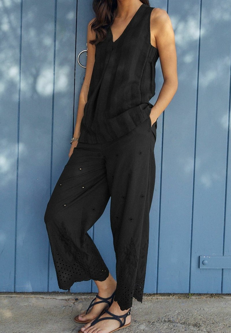 Femme portant un haut noir sans manches et un pantalon noir à jambes larges avec des motifs découpés, debout pieds nus devant une porte en bois bleue.