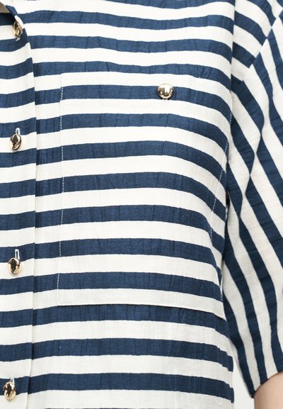 Μπλε ναυτικό και λευκό πουκάμισο με οριζόντιες ρίγες, χρυσά κουμπιά και τσέπη στο μπροστινό μέρος στο ύψος του στήθους, φορεμένο από άτομο με μανίκια τριών τετάρτων.