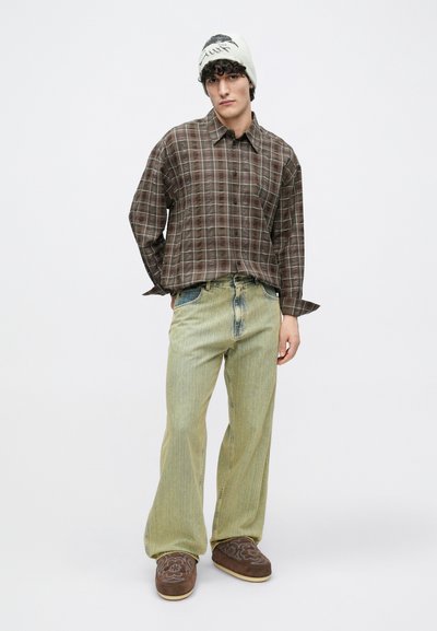 Brązowa koszula w kratę na guziki, jasne zielone spodnie o szerokich nogawkach oraz brązowe kapcie z kwiatowym haftem; na głowie kremowa czapka z czarnym wzorem.