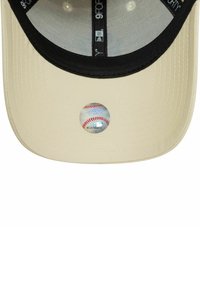 Gorra de béisbol color crema con visera negra, que presenta un gráfico de béisbol en el frontal y una tela texturizada con relieve.