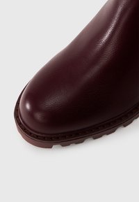 Bordo ādas potītes zābaks ar apaļu purngalu un teksturētu virsmu, kam ir bieza gumijas zole un redzamas šuves ap maliņu.