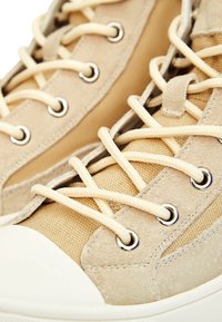 Béžové plátěné a semišové vysoké tenisky s oválnými očky, krémovými tkaničkami a bílým gumovým špičkovým krytem. Texturovaný povrch na semiši.