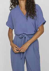 Top a maniche corte a V in morbido tessuto blu. Le maniche arrotolate e il punto vita annodato creano una silhouette rilassata ma aderente.