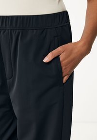 Svarta träningsbyxor med resårmidja och sidofickor, visade med en hand i fickan, vilket framhäver deras släta textur.