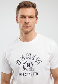 Baltas kokvilnas T-krekls ar drukātu "DENIM" logo tumši zilā krāsā, ar apļa emblēmu un teksta akcentiem zem tā. Īsas piedurknes un apaļa kakla izgriezums.