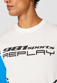 White cotton t-shirt with blue and black accents, featuring the text "981sports REPLAY" in bold black font across the front.
