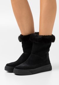 Jambes portant des bottes mi-mollet en daim noir avec des semelles en caoutchouc épaisses et une bordure en fourrure douce autour du haut sur un fond blanc.