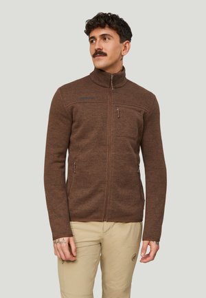 Mand med mørkt, krøllet hår og overskæg iført en brun fleecejakke med lynlås og beige bukser, der kigger let til siden.