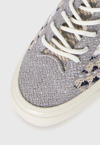 Zapatilla de punto gris con cordones crema, parte superior de tela estampada y suela de goma blanca. Presenta detalles texturizados y punta redonda.