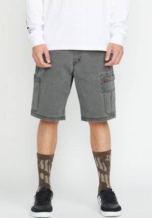 Grijze cargoshorts met twee grote zijzakken en contrasterende stiksels. Gecombineerd met gemusterde bruin en beige sokken en zwarte sneakers.