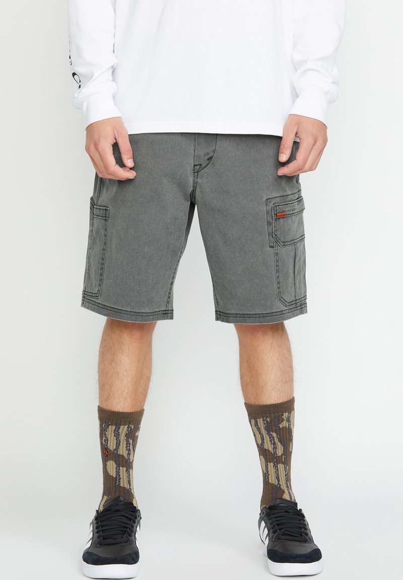 Graue Cargo-Shorts mit zwei großen Seitentaschen und kontrastierenden Nähten. Kombiniert mit gemusterten braunen und beigen Socken sowie schwarzen Sneakers.