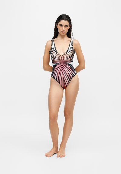 Femme debout, pieds nus, portant un maillot de bain une pièce avec un motif rayé symétrique noir et rose, sur un fond uni clair.