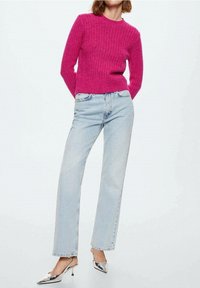 Vrouw in een felroze gebreide trui, lichtblauwe rechte jeans en zilveren slingback hakken met puntige neus, staande met haar handen achter haar rug.