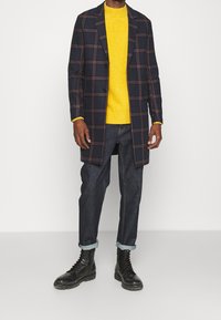 Marine-karierter langer Mantel über einem gelben Strickpullover, kombiniert mit dunklen Jeans und schwarzen Schnürstiefeln. Der Mantel hat zwei Knöpfe und Reverskragen.