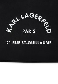 Melna auduma soma ar baltu tekstu: "KARL LAGERFELD," "PARIS," un "21 RUE ST-GUILLAUME" drosmīgā, izliekta dizainā.