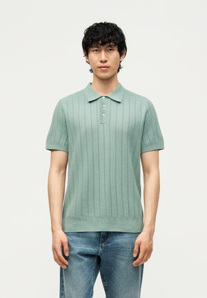 Fiatal férfi göndör fekete hajjal, aki világoszöld, rövid ujjú, bordázott galléros ingben és kék farmerral áll egyszerű háttér előtt.