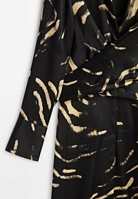 Schwarzes Oberteil aus Stoff mit goldenen Pinselstrichmuster. Besitzt lange Ärmel und ein Wickeldesign, glatte Textur mit leichtem Glanz.