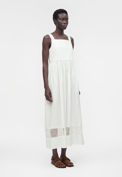 Γυναίκα με σκουρόχρωμο δέρμα που φοράει λευκό φόρεμα μίντι χωρίς μανίκια με δαντελένια λεπτομέρεια κοντά στο τελείωμα και καφέ φλατ σανδάλια, στέκεται μπροστά σε λευκό φόντο.