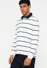 Polo cu mâneci lungi, de culoare albă, cu dungi orizontale în nuanțe de bleumarin și albastru deschis. Dispune de un guler bleumarin și un logo pe partea stângă a pieptului.