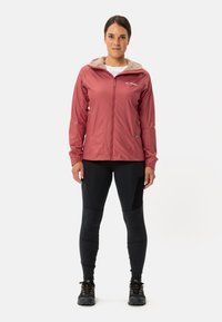 Rote wasserdichte Jacke mit Reißverschluss und beigem Kapuzenfutter, kombiniert mit schwarzen Leggings und Wanderschuhen. Mit einem Logo auf der Brust.